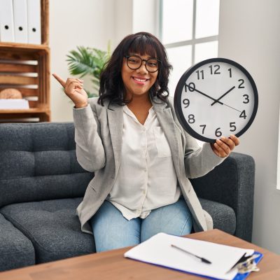 Hispanic woman working at therapy office holding clock smiling happy pointing with hand and finger to the side Hispanic woman working at therapy office holding clock smiling happy pointing with hand and finger to the side